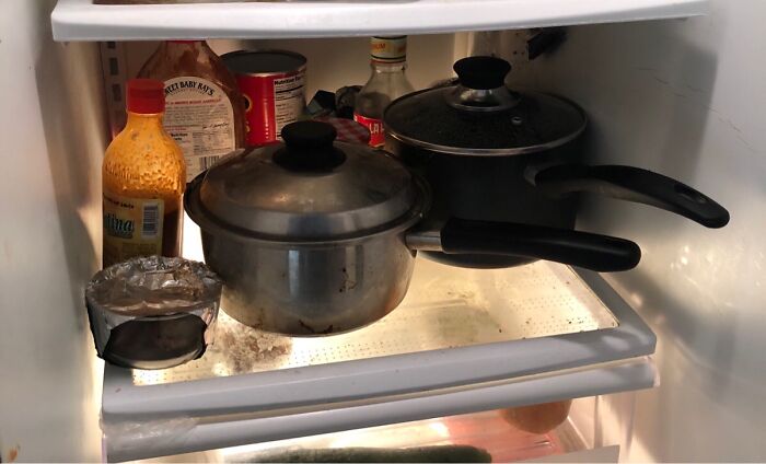 A dirty refrigerator shelf with pots, bottles, and a foil-covered dish, depicting a horrible roommate situation.