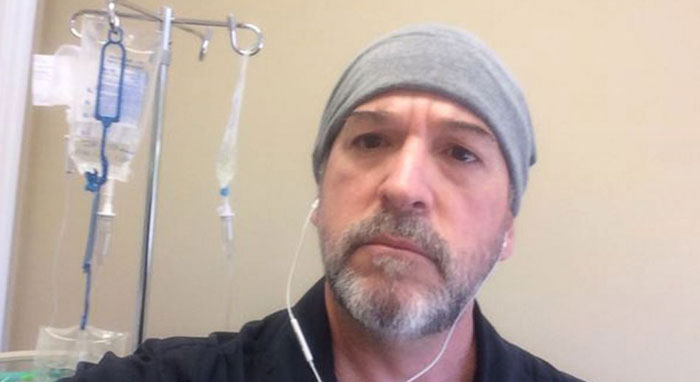 Man wearing beanie and earbuds in hospital setting with IV bags in background
