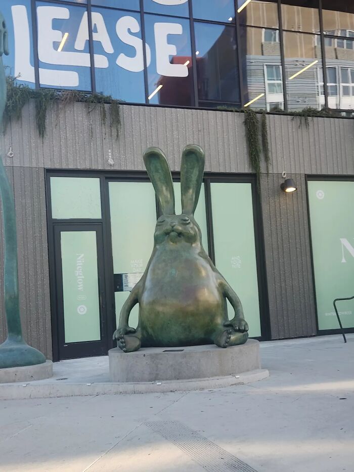 A bizarre, large, green bronze statue of a contemplative rabbit with long ears, sitting outdoors.