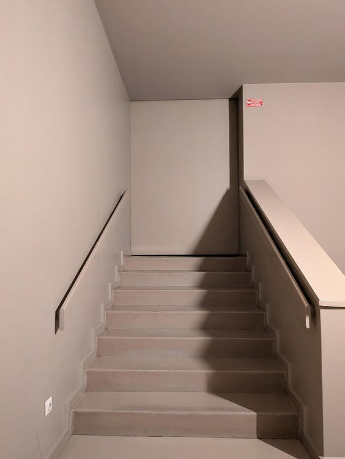 Upward view of an unsettling, empty liminal space staircase with beige walls and a dark handrail.