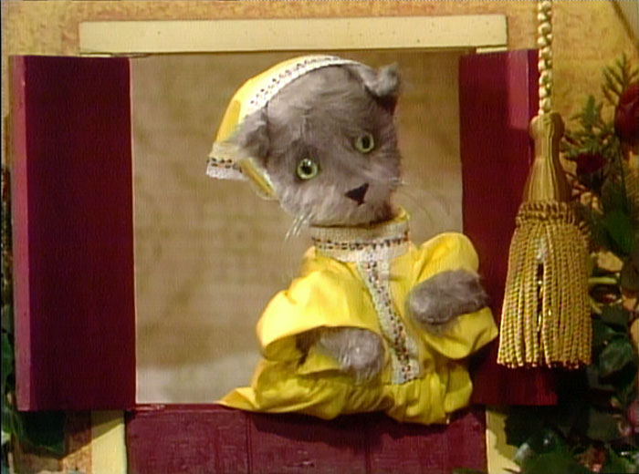A puppet cat in a yellow dress and bonnet looks out a window, a nod to the sister's meow comeback. A puppet cat in a yellow dress and bonnet looks out a window, a nod to the sister's meow comeback.