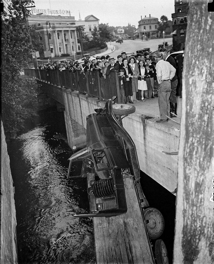 Black and white historic photo of a car accident with a vehicle hanging off a bridge and crowd gathered
