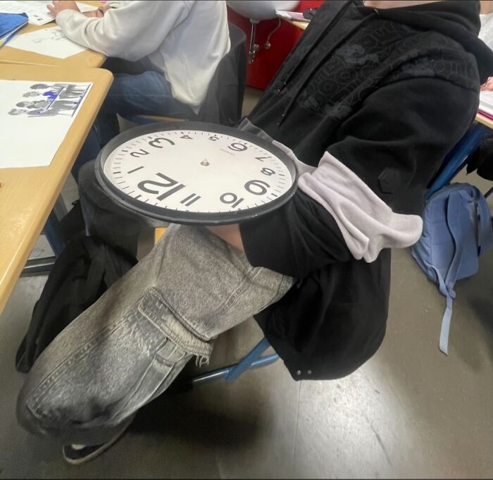 A person holding a large, bizarre clock face without hands, while seated in a classroom.