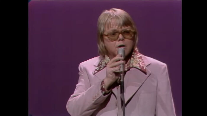 A male singer with a microphone and sunglasses performing on stage, taking you way back and reminding you how old you are.
