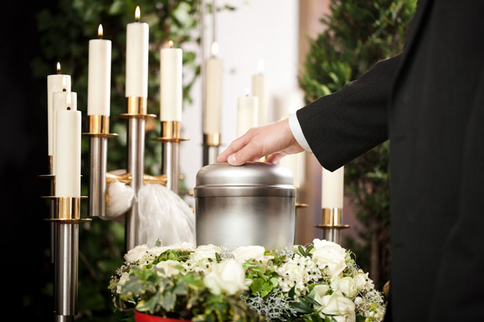 Hand touching silver urn surrounded by white flowers and candles at memorial service Hand touching silver urn surrounded by white flowers and candles at memorial service