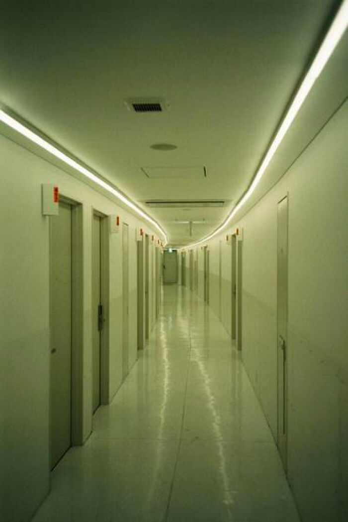 A long, fluorescent-lit liminal space in a curved hallway with numerous doors, evoking a haunting feeling.