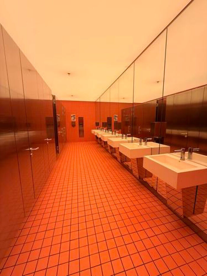 A restroom with vibrant orange tiles, long mirrors, and multiple sinks, an unsettling haunting liminal space.