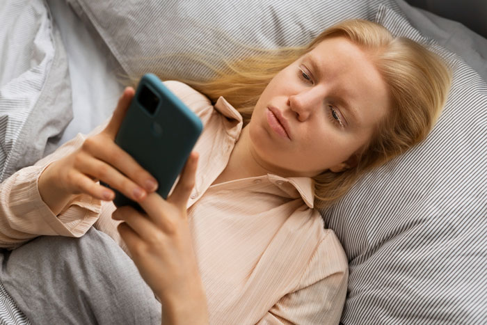 A woman, lying in bed under striped covers, looking at her phone, reflecting on her talkative fiancé situation. A woman, lying in bed under striped covers, looking at her phone, reflecting on her talkative fiancé situation.