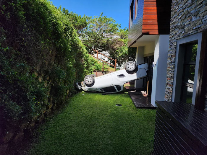 An overturned white car, a delivery fail, lies on a grassy lawn next to a modern house and lush green hedge.