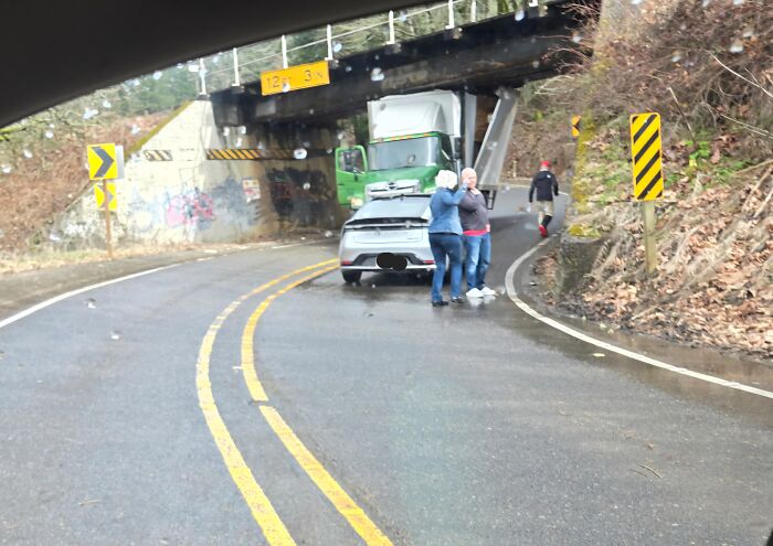 A truck stuck under a low bridge, blocking the road, with people standing around it. A silly solution.
