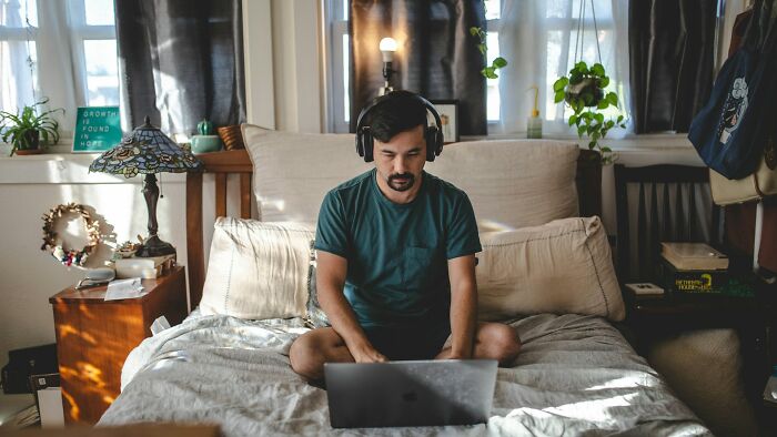 A man with headphones working on a laptop in bed, depicting potential intense stress and burnout from work.