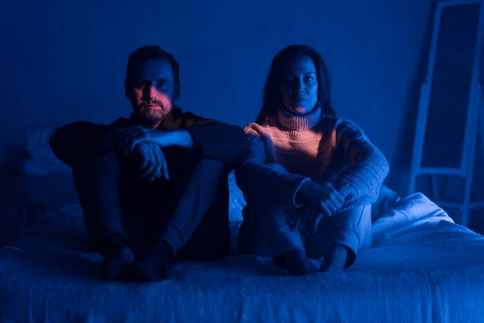 A man and woman sit separated on a bed in dim light, depicting the emotional distance of a marriage ending.