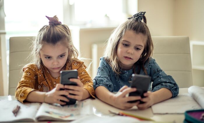 Two young girls distracted by phones while sitting at table with books, showing Gen Alpha illiteracy concerns