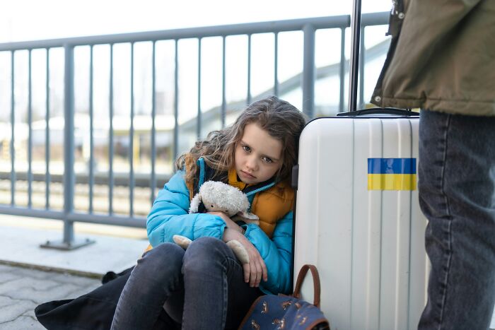 Sad girl with teddy bear sitting by suitcase on street showing family secrets impact