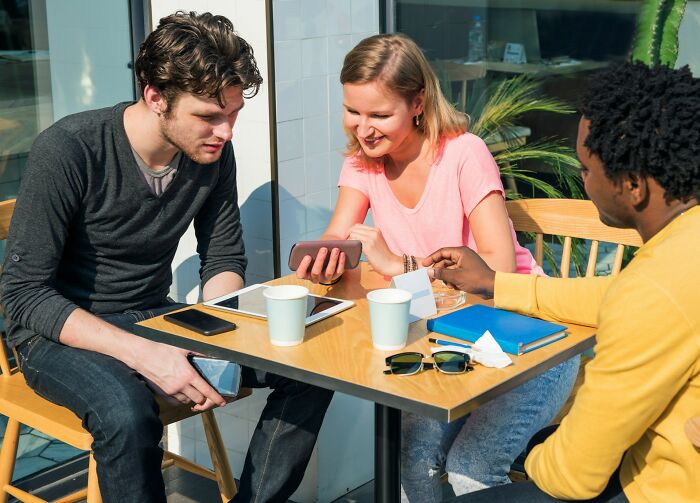 Friends chatting and showing phone at outdoor cafe table with coffee cups