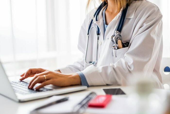 A female doctor in a white coat typing on a laptop, symbolizing intense stress and burnout among healthcare professionals.