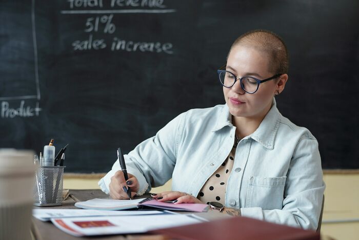 Student writing notes in classroom with chalkboard behind her showing school content