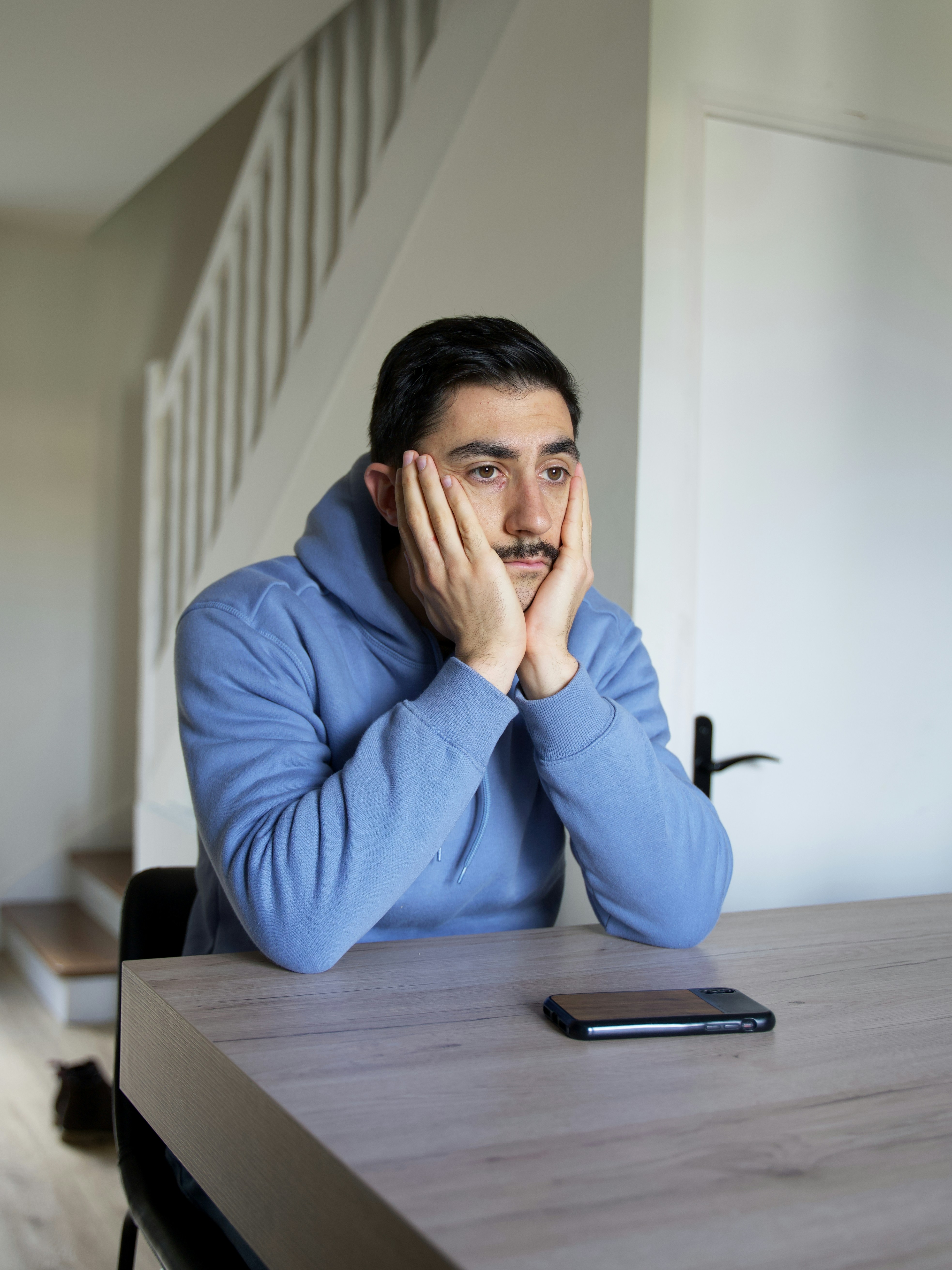 A husband with a mustache and blue hoodie sits at a table looking dejected, feeling blind-sided by the family trip.