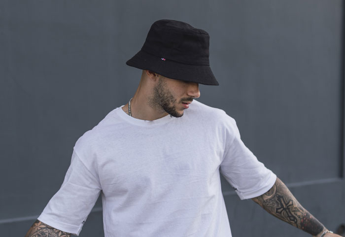A man wearing a black bucket hat and white t-shirt, looking down. People tell their partners everything.