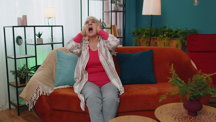 An older woman with a breakdown, screaming and holding her head, sitting on a sofa in a living room.