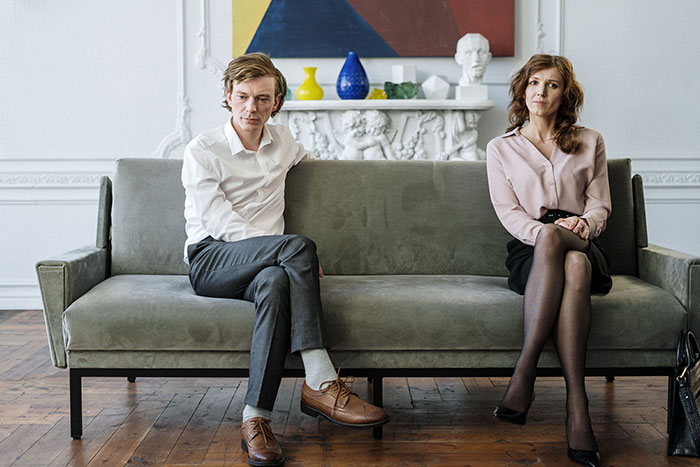 A man and woman sitting separately on a couch, looking distressed, depicting a husband breaking his promise of a postnup after years of marriage. A man and woman sitting separately on a couch, looking distressed, depicting a husband breaking his promise of a postnup after years of marriage.