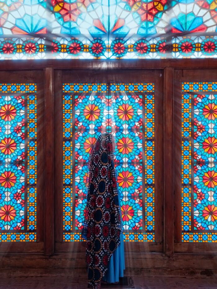 Person wearing patterned clothing in front of Persian stained glass window with floral geometry