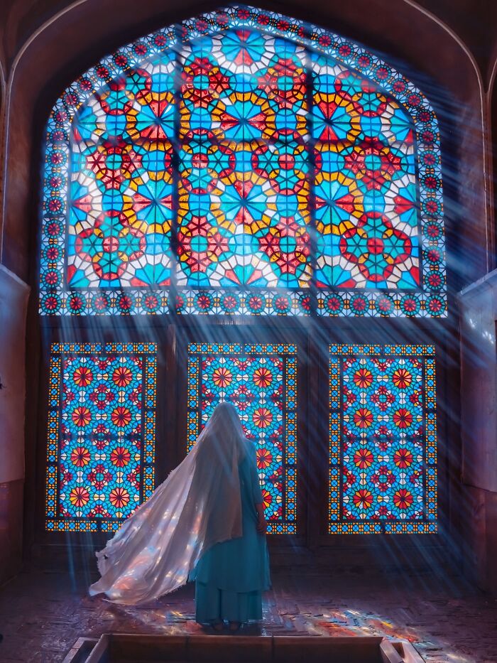Person in traditional clothing stands before colorful Persian stained glass window with geometric patterns