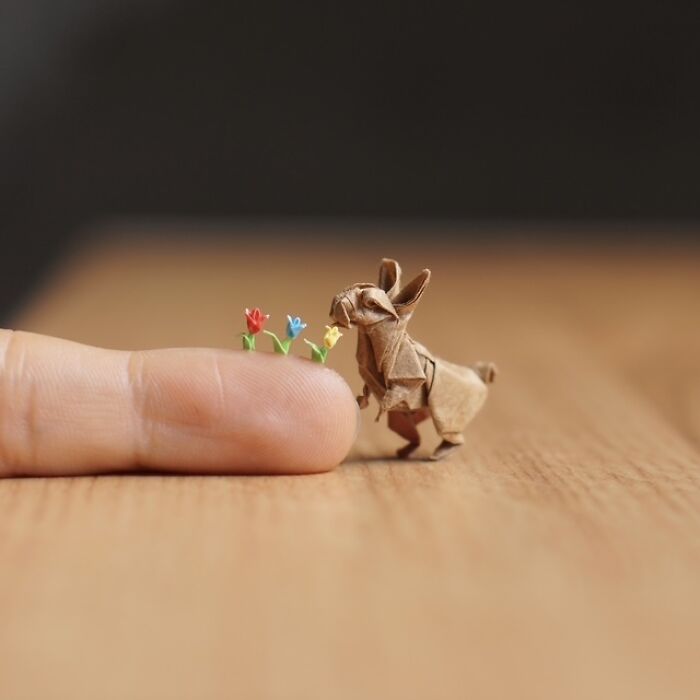 Miniature paper figure of a rabbit sniffing tiny flowers on a finger, showcasing immense detail.