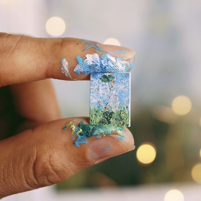 Artist creates miniature paper figures, a tiny replica of a painting, held by paint-stained fingers with immense detail.