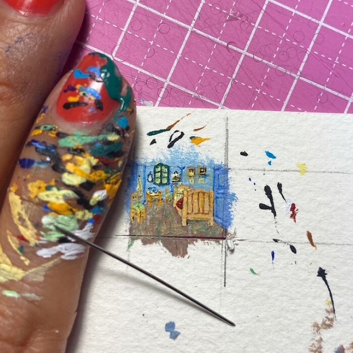 A heavily paint-stained finger and needle next to a detailed miniature paper figure painting of a bedroom interior.