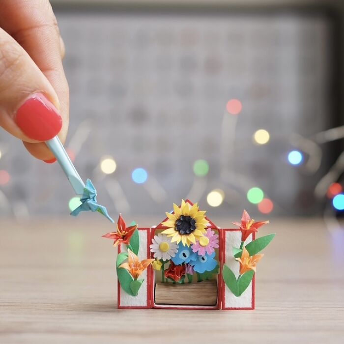 A hand holding a tiny paper figure of a bird near a miniature paper flower arrangement, showcasing immense detail.