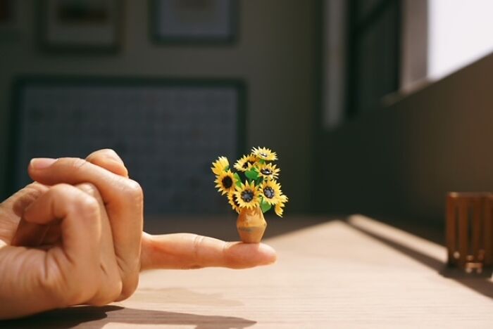 A hand balancing a miniature vase filled with tiny paper sunflowers, demonstrating immense detail.
