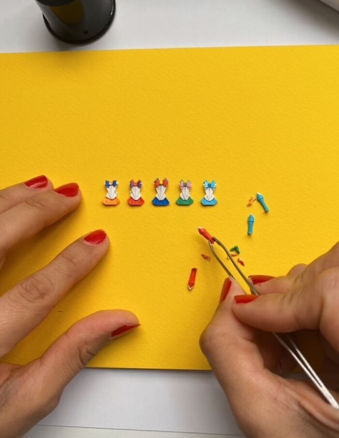 Hands using tweezers to craft miniature paper figures on a yellow surface, highlighting their intricate detail.