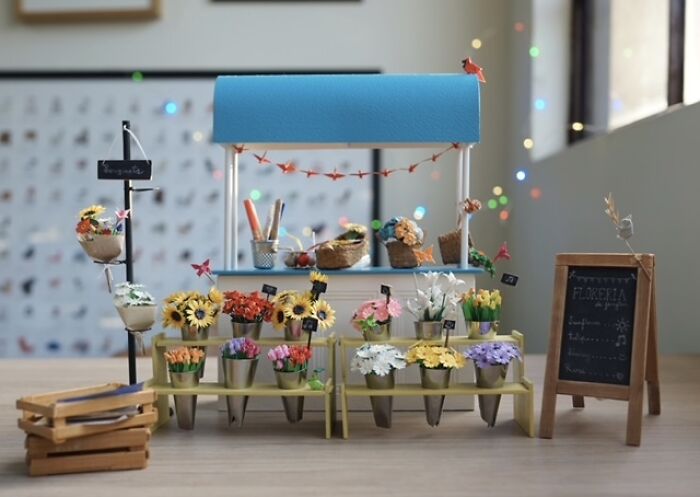 A detailed miniature paper figure of a flower shop with tiny potted plants and a blue awning.