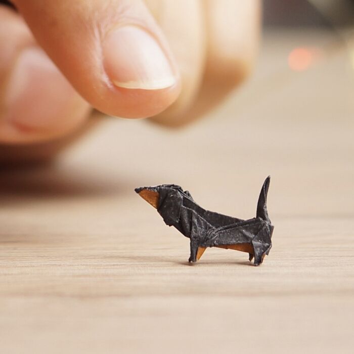 Artist creates miniature paper figures, a black paper dog on a wooden surface, with a finger approaching, showing immense detail.