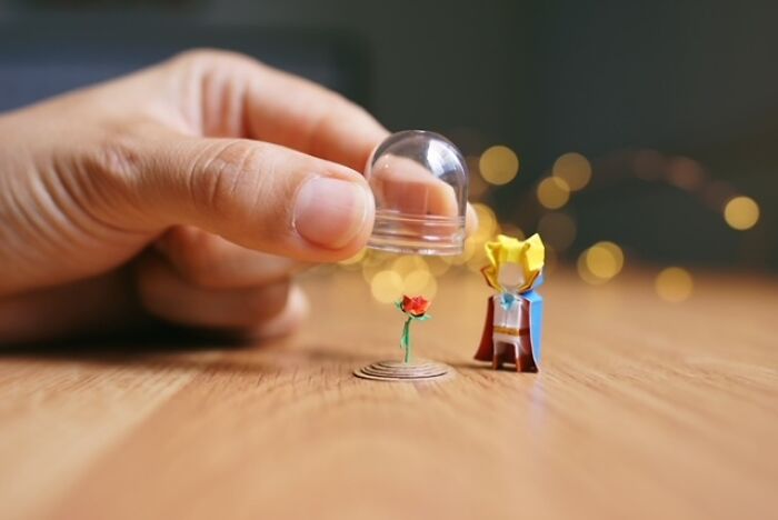 A human hand holds a clear dome over a miniature paper figure of a rose next to The Little Prince.