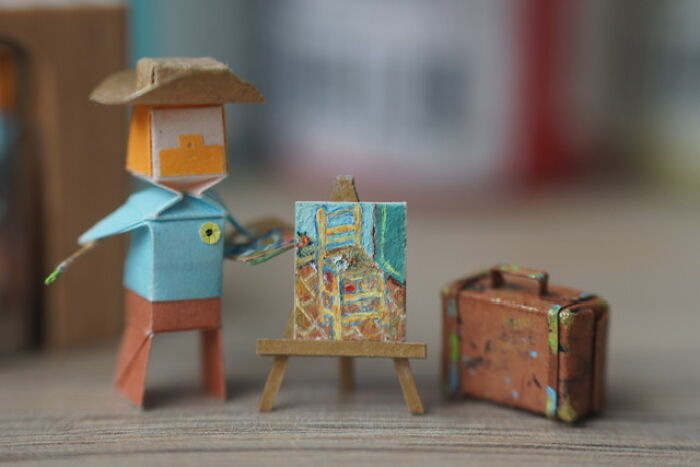 A tiny paper figure of an artist in a hat painting on a miniature easel next to a small suitcase.