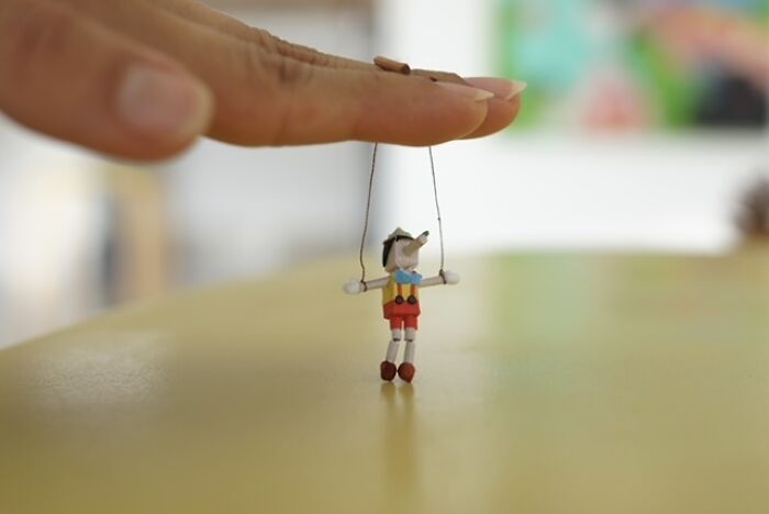 A human hand manipulates a miniature paper figure of Pinocchio, hanging by strings like a marionette.