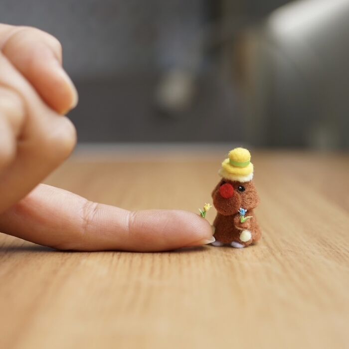 Miniature paper figure wearing a hat and holding flowers, presented with immense detail on a finger.