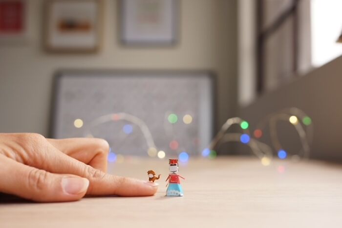 A human finger points at a small paper figure of a nutcracker soldier and a tiny dog, with bokeh lights.