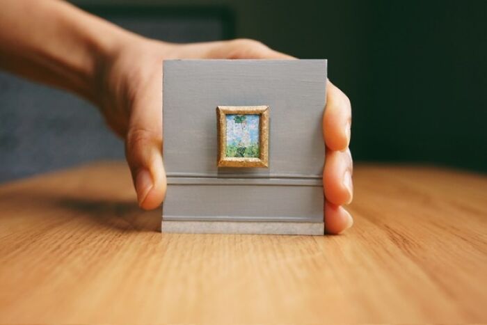 A hand holding a miniature paper figure of a framed landscape painting, showcasing immense detail in the tiny artwork.