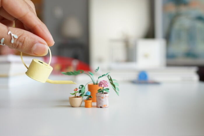 Hand watering miniature paper figures of potted plants with a tiny yellow watering can.
