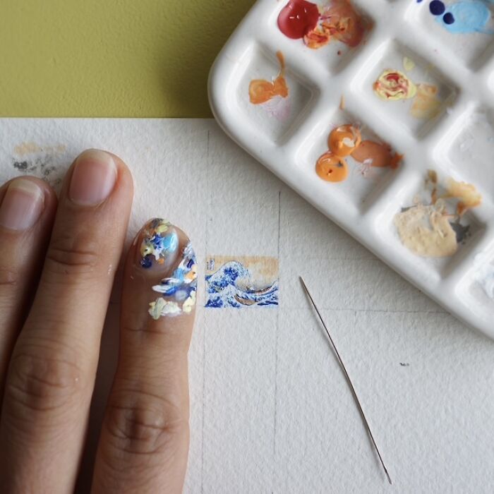 Artist creating a miniature paper figure of a wave on a tiny canvas with paints and a needle.