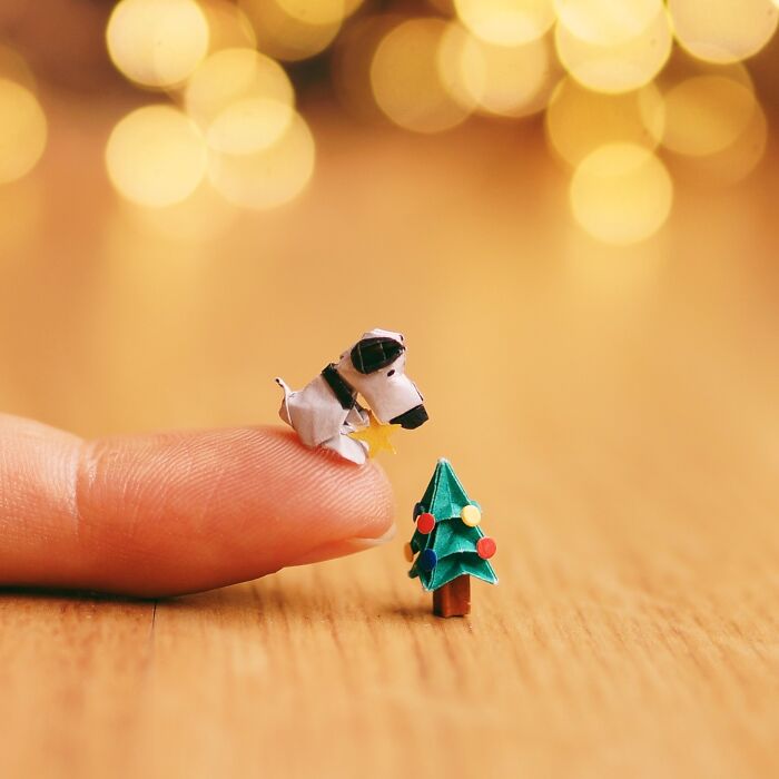 Artist creates miniature paper figures, a tiny paper dog on a finger admiring a miniature Christmas tree with immense detail.