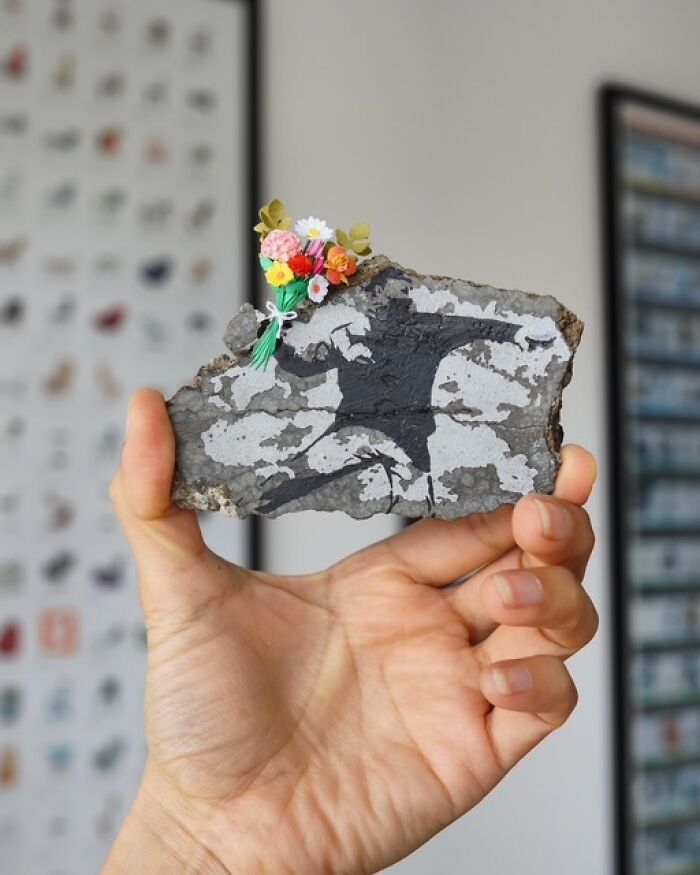 Miniature paper figure of a person holding a tiny bouquet of flowers, displayed on a piece of rock.
