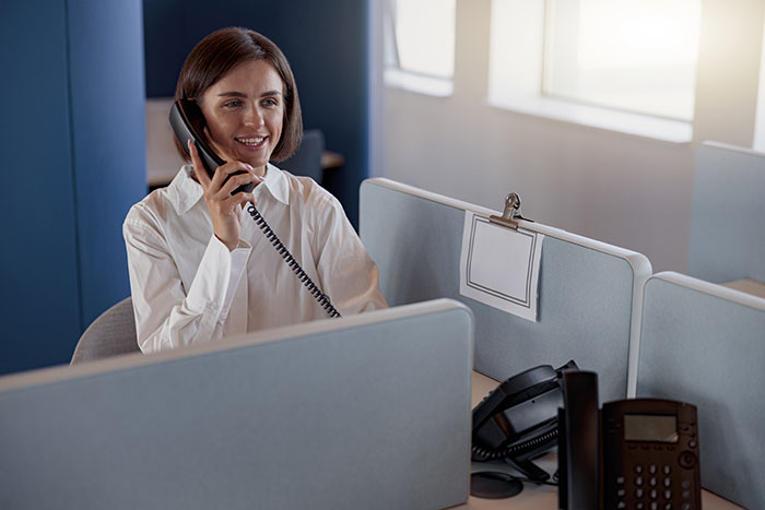 Smiling woman talking on a landline phone at an office desk, keeping industry secrets from customers.