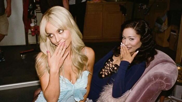 Two women smiling and covering mouths sitting on pink chair behind the scenes Two women smiling and covering mouths sitting on pink chair behind the scenes