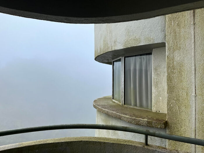 A curved concrete balcony and window of an abandoned building shrouded in fog create an unsettling liminal space. The scene descends into the abyss.