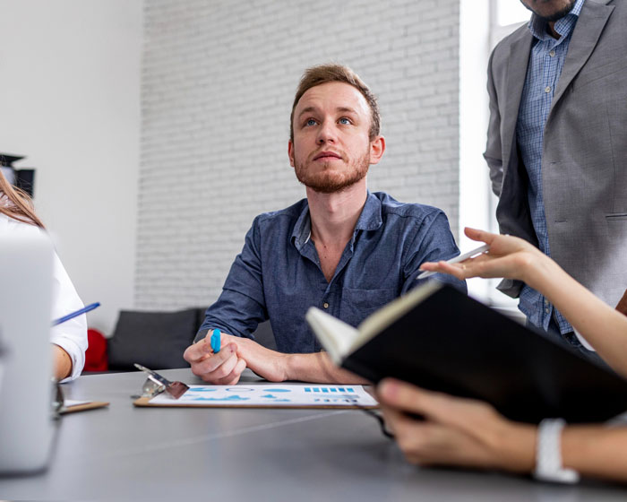 A man unjustly fired, sitting at a table with colleagues, looking up thoughtfully during a meeting.
