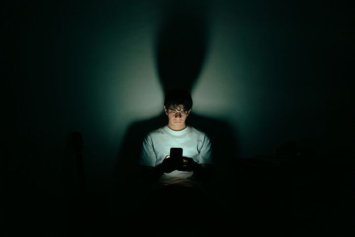 A person is using a mobile phone in a dark room. The phone's light illuminates their face, creating a shadow behind them. Learn useless facts.